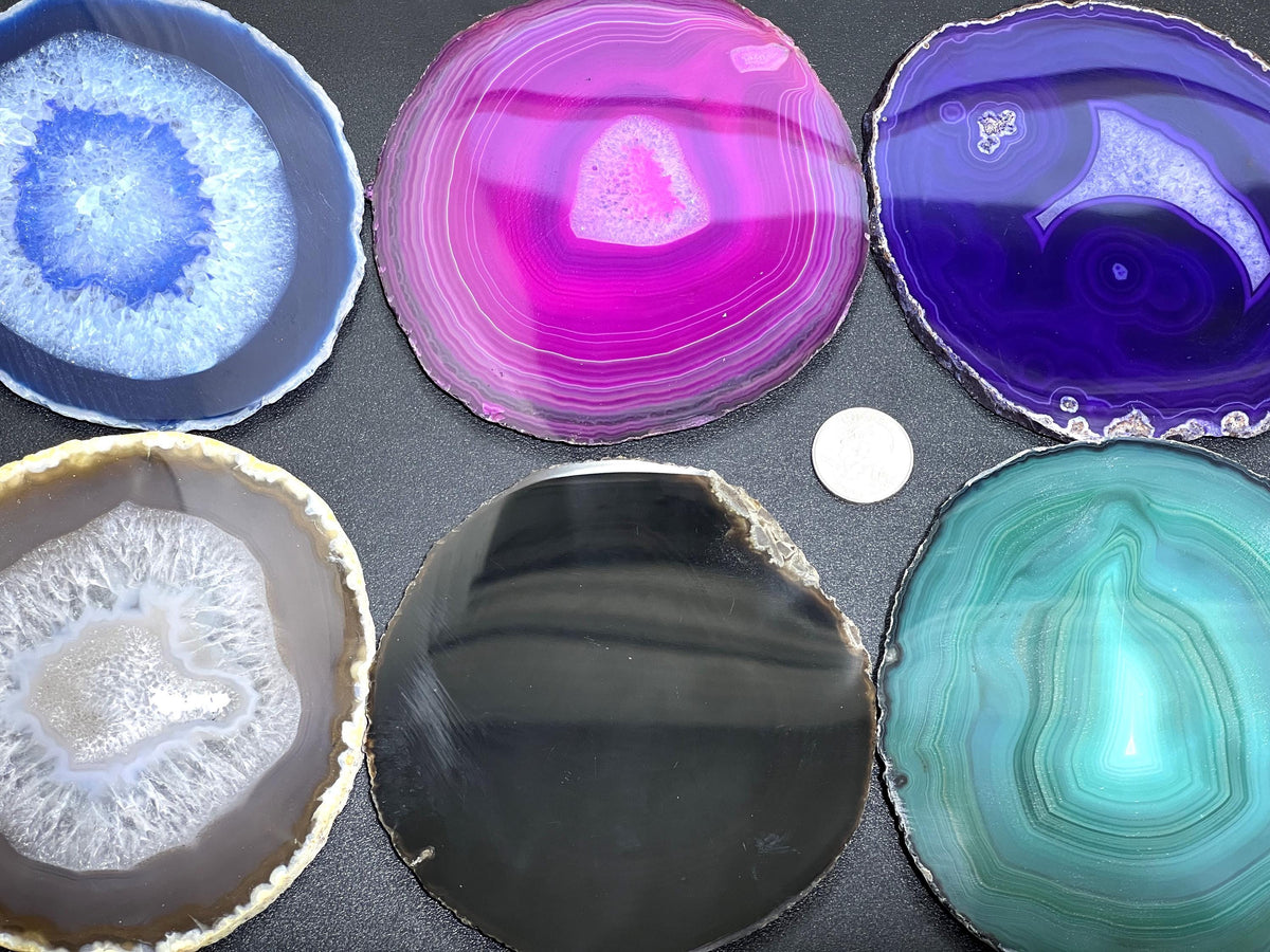 Assorted colorful polished agate slices on a dark surface beside a small round white size reference