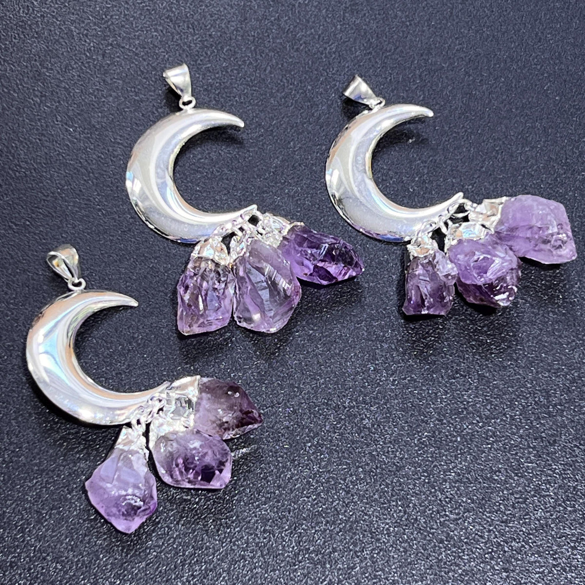 Three silver-plated crescent moon pendants, each with three dangling amethyst crystals, on a dark textured surface.
