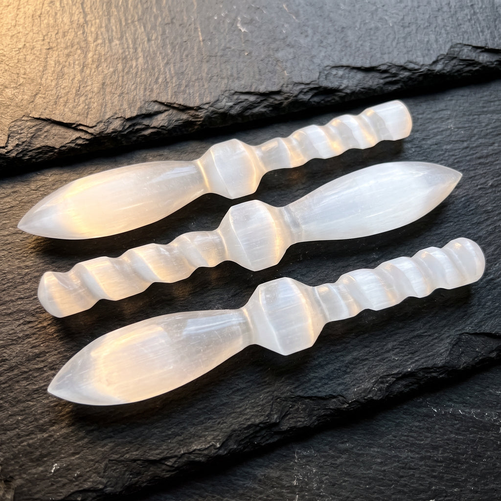 Three white satin spar decorative crystal daggers with spiral handles arranged on dark slate