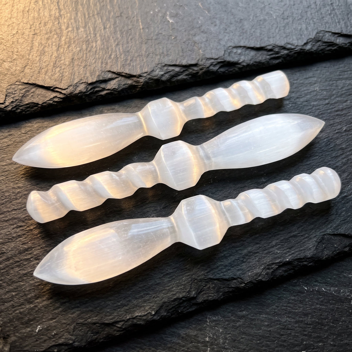 Three white satin spar decorative crystal daggers with spiral handles arranged on dark slate