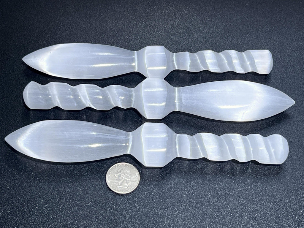Three selenite satin spar crystal daggers with spiral handles on a dark surface beside a coin for scale