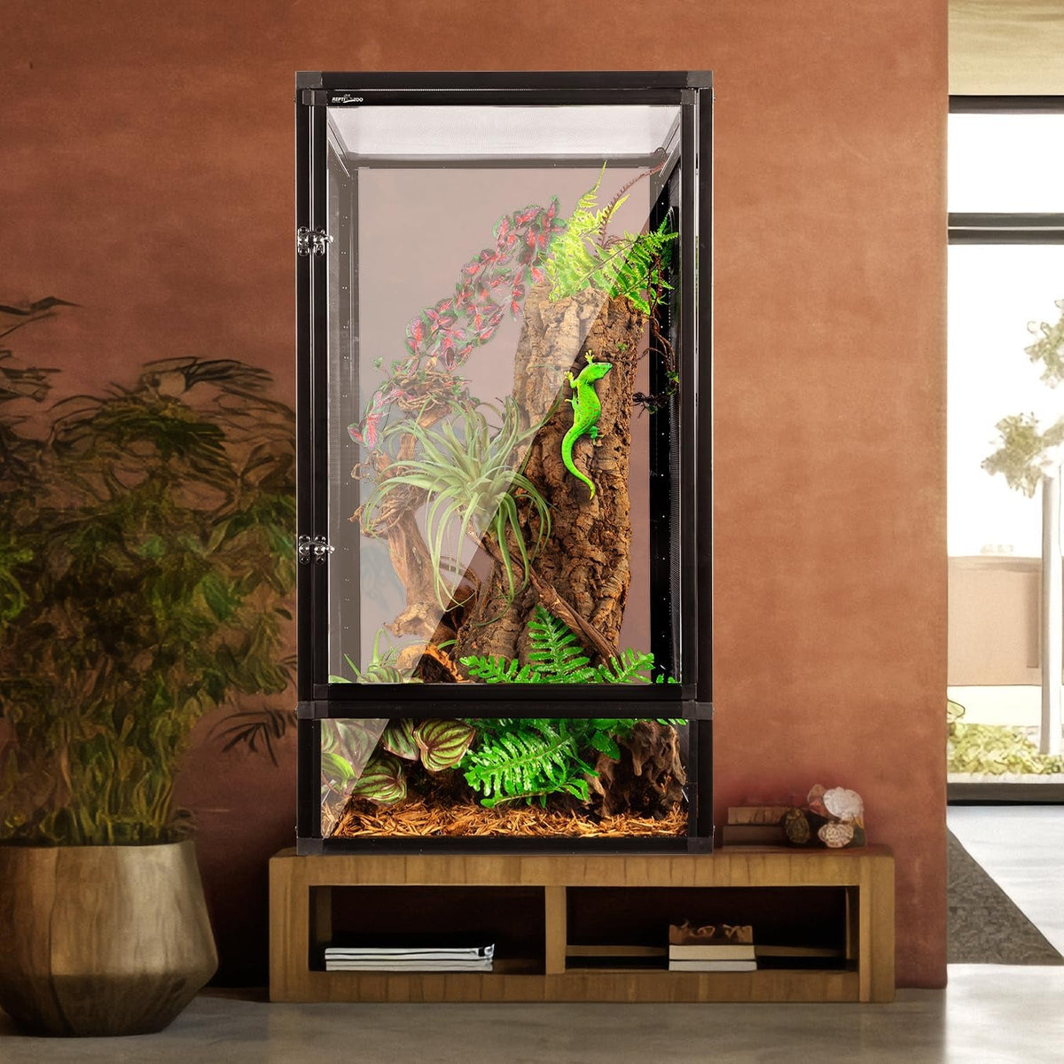 A tall screen terrarium with a black frame and interior climbing branches, plants, and a bright green gecko, displayed on a wooden stand against a terracotta-colored wall in a modern room setting.