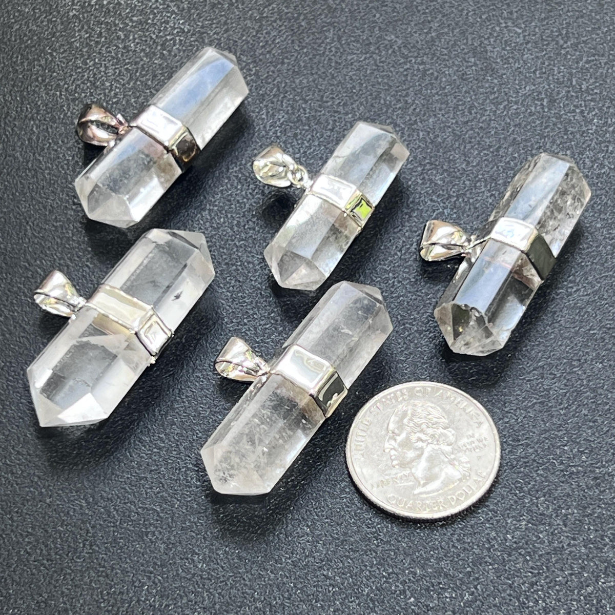 Five double-pointed quartz pendants with silver-plated fittings beside a quarter for size comparison.
