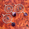 Three silver-plated dreamcatcher pendants with purple amethyst centers and hanging crystals on reddish wood