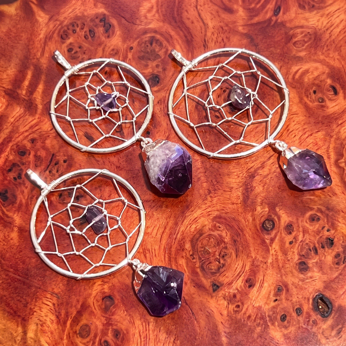 Three silver-plated dreamcatcher pendants with purple amethyst centers and hanging crystals on reddish wood