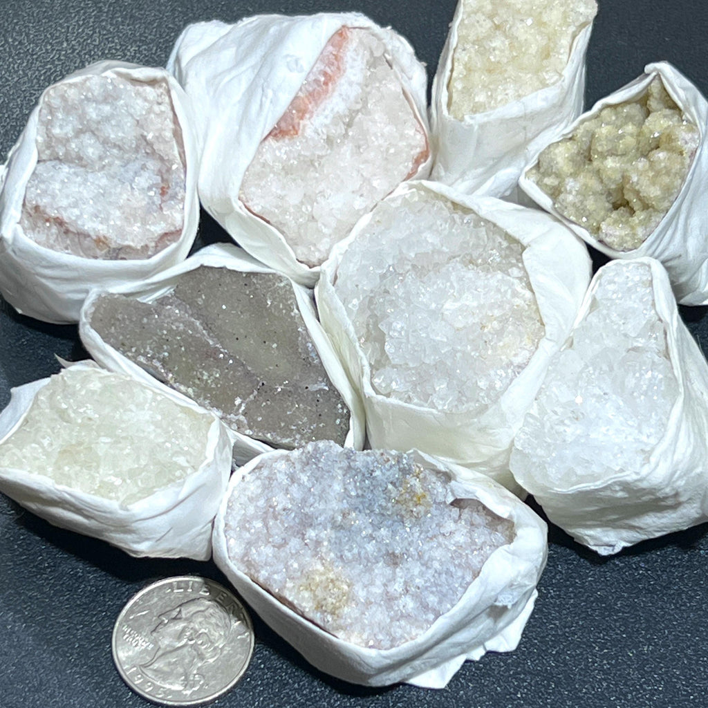 Mixed druzy crystal clusters wrapped in white paper on a dark surface, with a quarter for scale