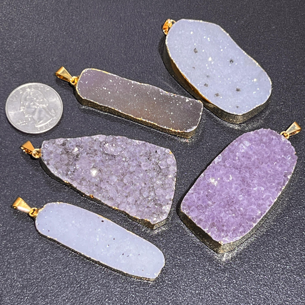 Five assorted druzy pendants with gold-colored rims on a dark surface beside a quarter for scale