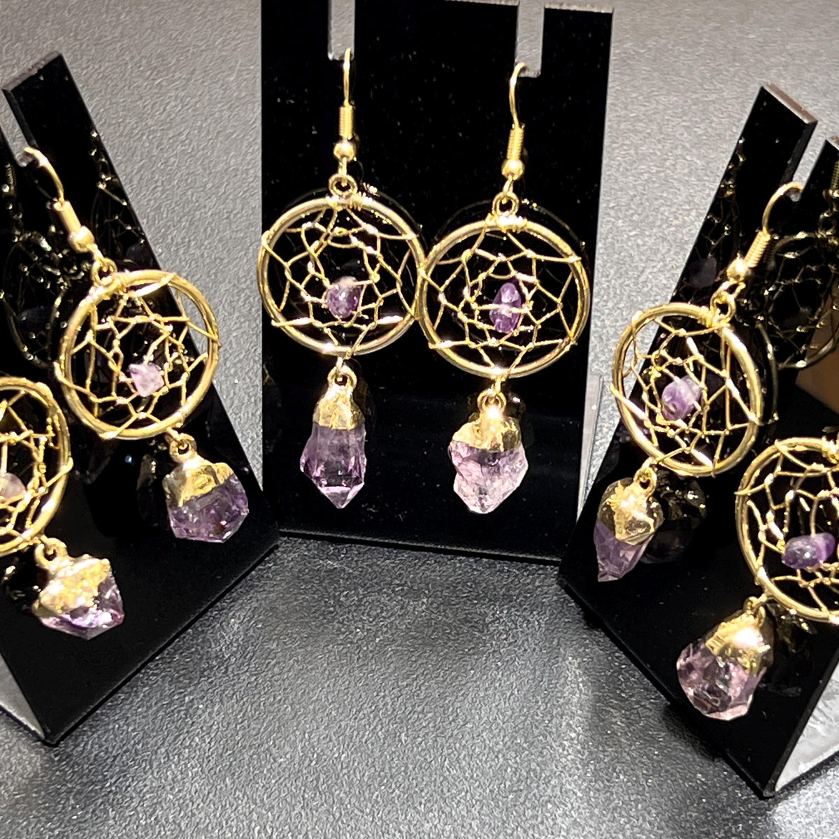 Close-up of gold-plated dreamcatcher earrings with small purple center crystals and dangling amethyst points on black stands.

