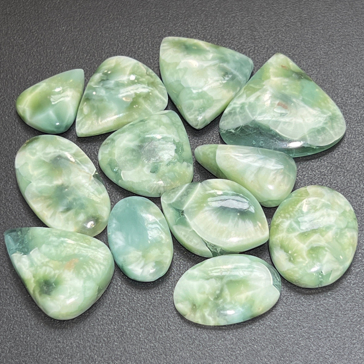 Assorted green and blue-green hemimorphite cabochons arranged on a dark surface, including oval, teardrop and rounded triangular shapes