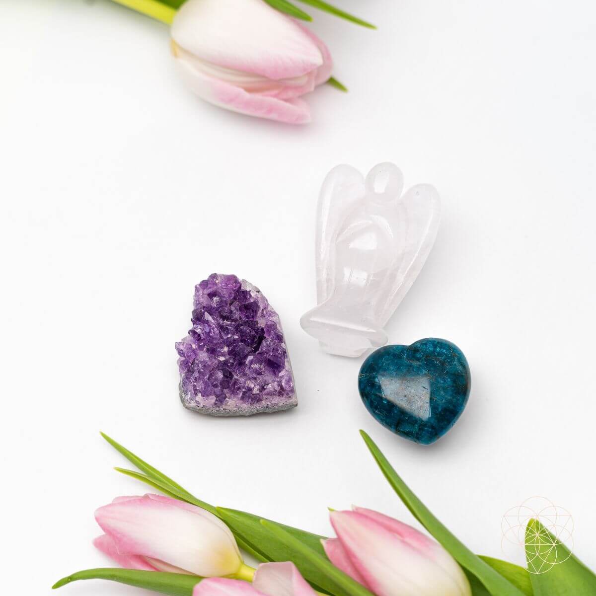Clear Quartz angel, purple Amethyst cluster, and blue Apatite heart arranged on white beside pink tulips.