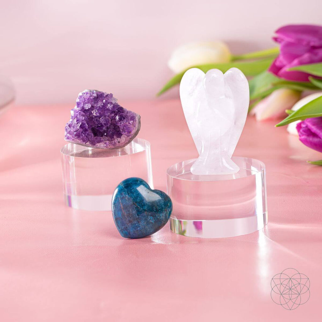 Amethyst cluster, Clear Quartz angel, and blue Apatite heart on clear display stands against a pink background.
