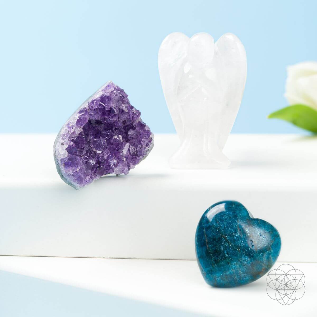 Blue Apatite heart in front of an Amethyst cluster and Clear Quartz angel on a white stepped display.