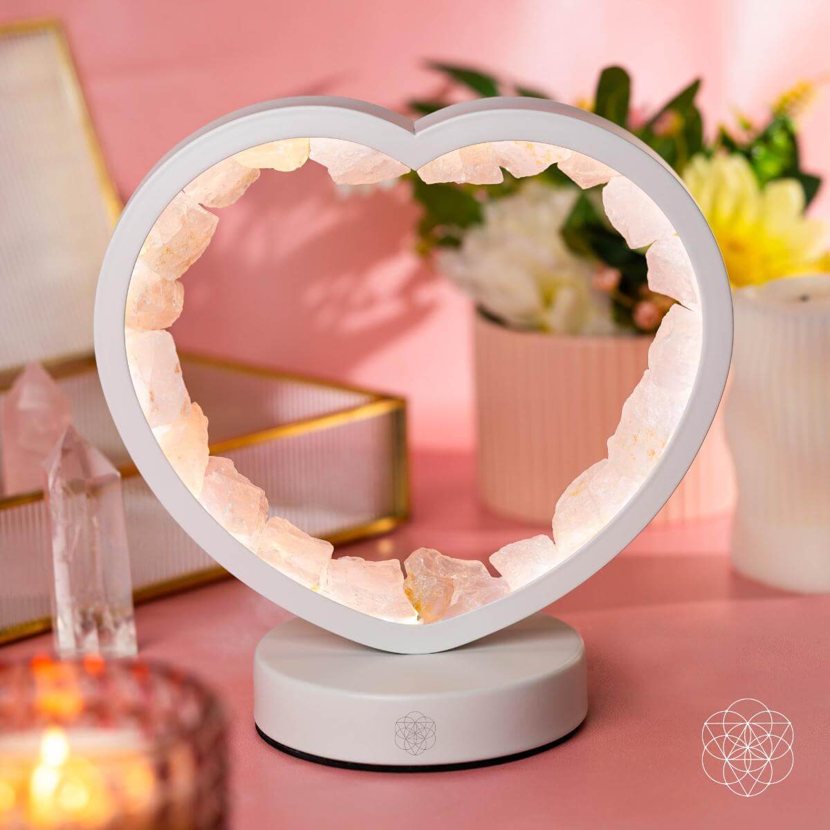 Pale heart-shaped rose quartz lamp glowing on a pink desktop beside flowers, a candle, and crystal points.
