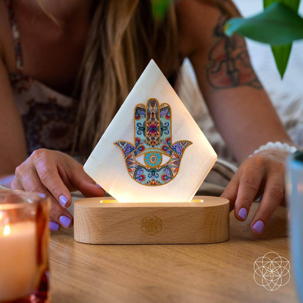 Person arranging an illuminated selenite Hamsa hand lamp on a wooden table beside a candle
