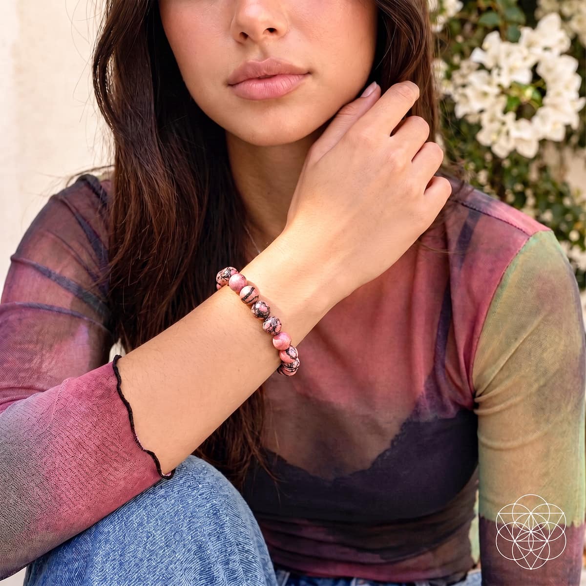 Woman wearing a pink-and-black rhodonite bead bracelet with a multicolored sheer top and jeans.
