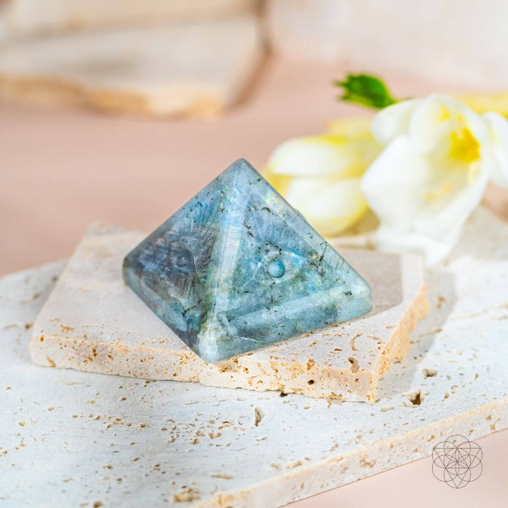 Carved labradorite eye pyramid with blue-green flashes on cream stone slabs beside white flowers.
