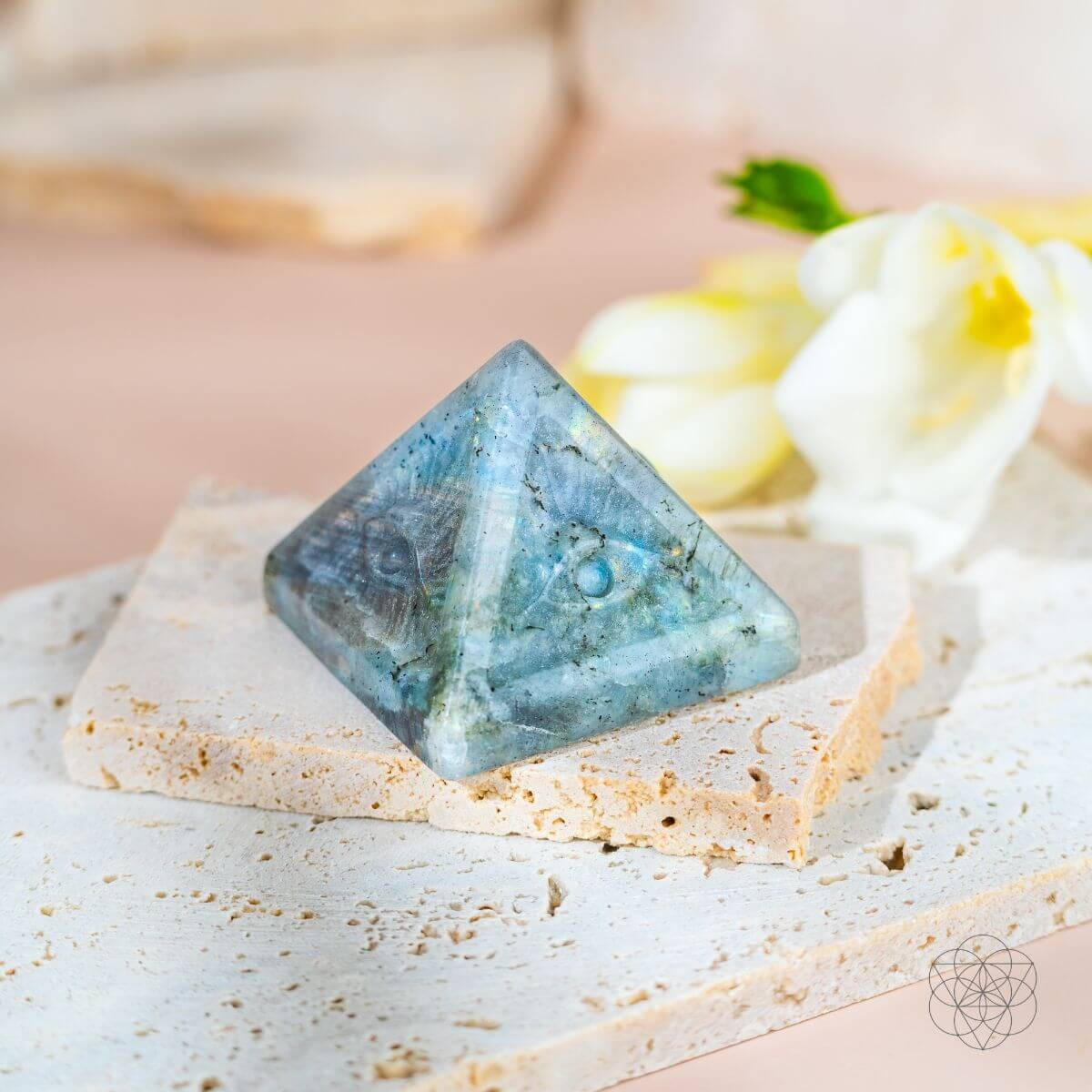 Carved labradorite eye pyramid with blue-green flashes on cream stone slabs beside white flowers.
