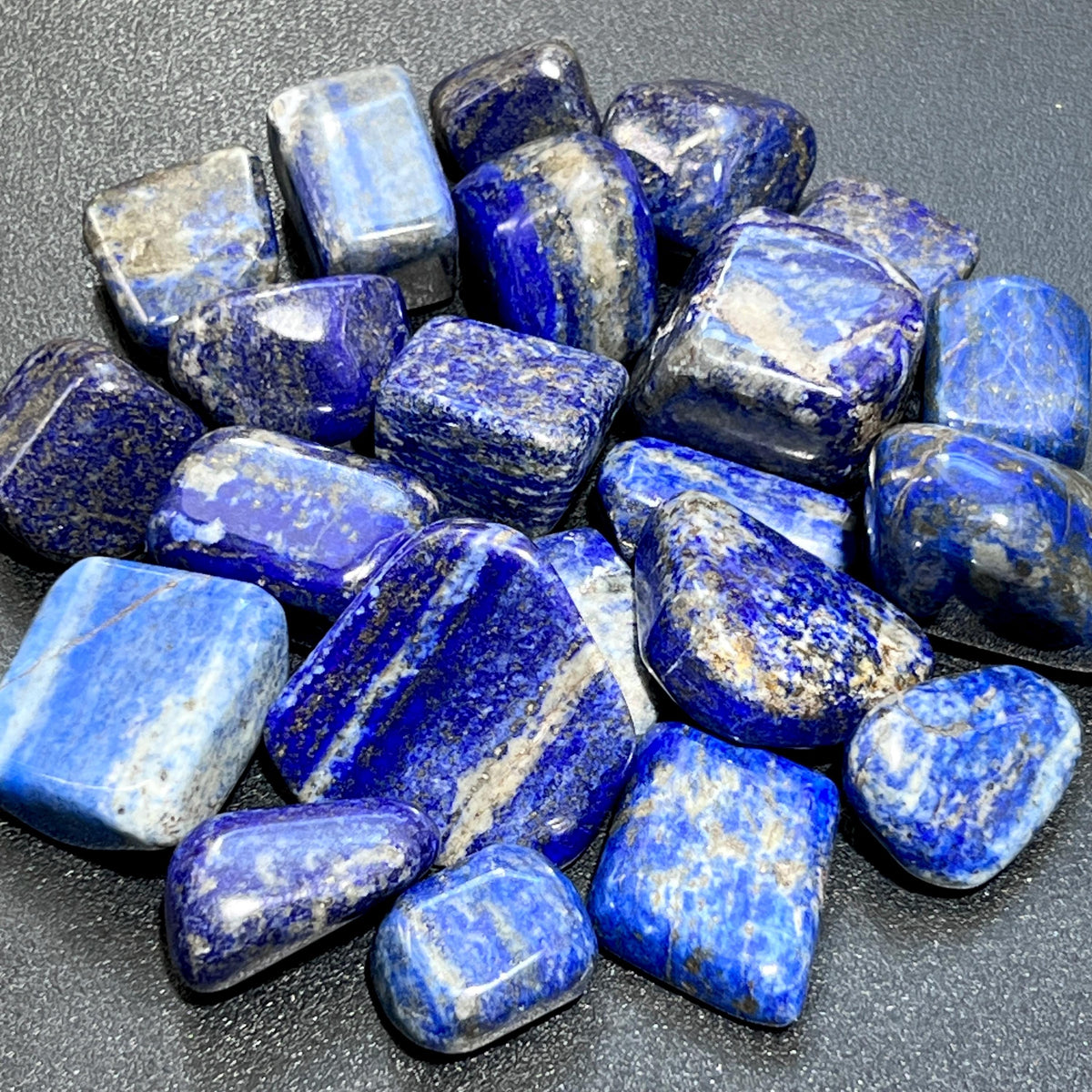 Assorted polished lapis lazuli stones with deep blue color, white and gray bands, and gold-colored speckles on a dark surface.

