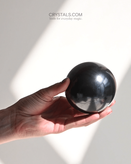 Hand holding a glossy black shungite sphere in sunlight against a pale background.

