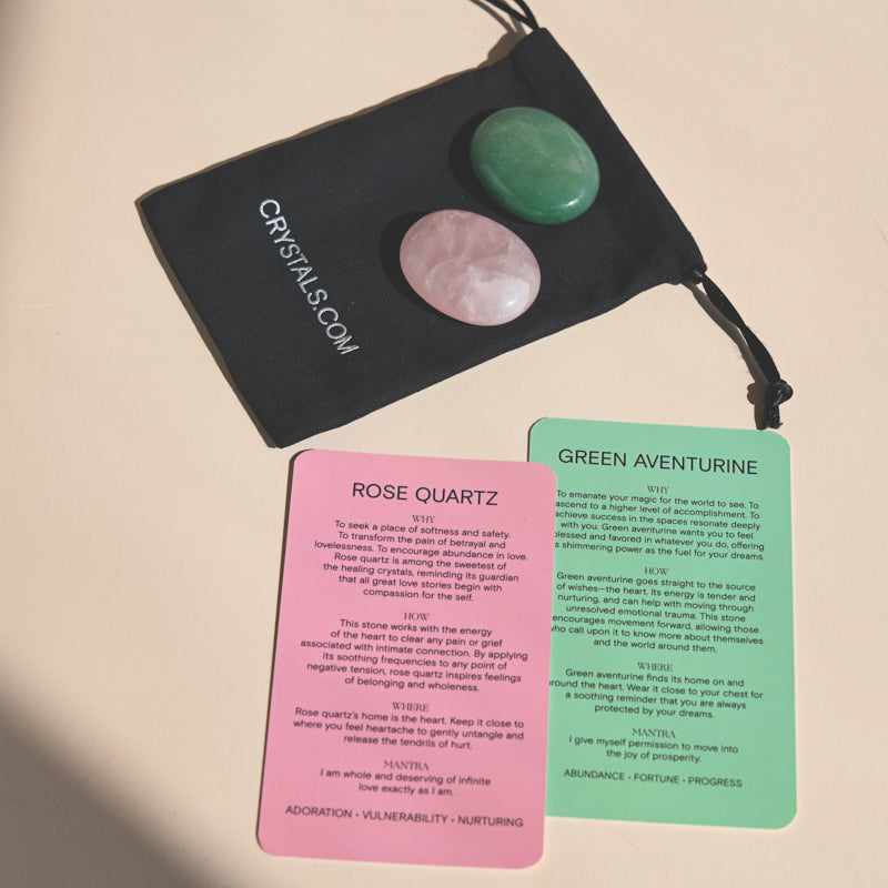 Two polished palm stones, pink rose quartz and green aventurine, displayed on a black drawstring pouch with printed cards.
