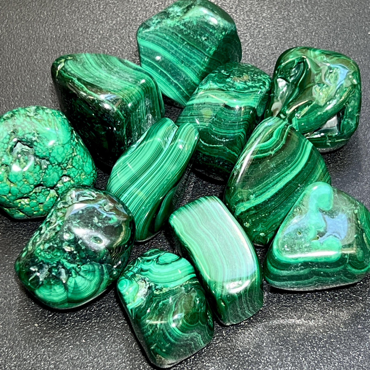 Large polished malachite tumbled stones with bright and dark green bands, curves, and ring patterns on a dark surface.