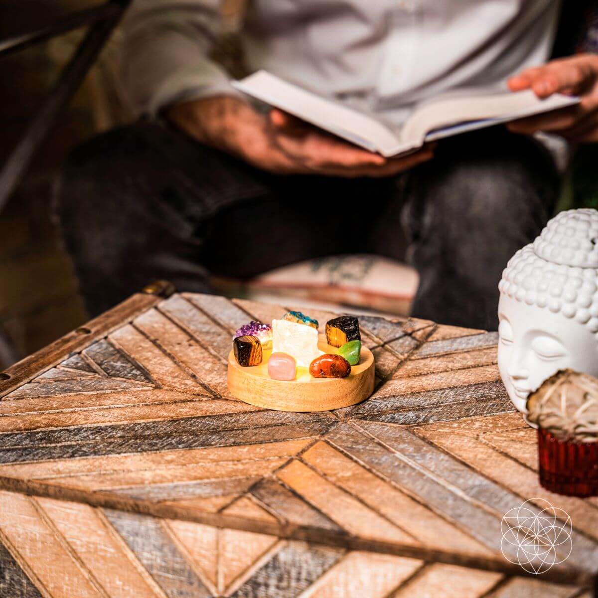 Illuminated mixed-crystal lamp on a patterned wooden table in front of a person reading a book.
