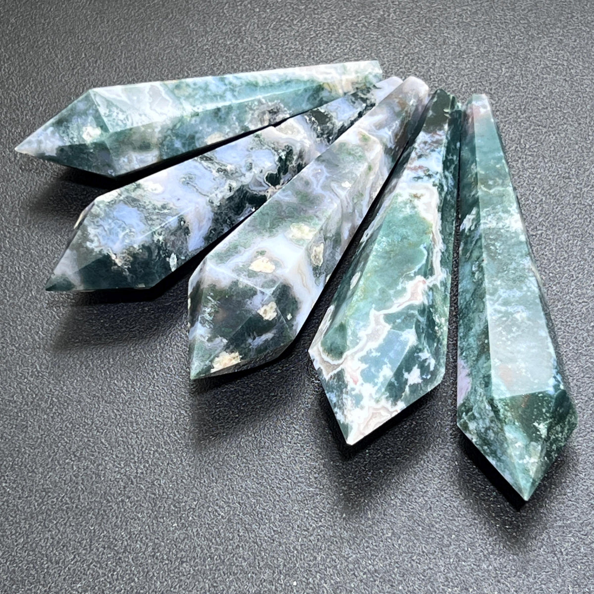 Five faceted moss agate scepter wands with green and white patterns arranged in a fan on a dark surface.
