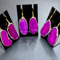  Three pairs of vivid pink agate slice earrings with gold-plated edges and gold-colored hooks on black display stands.