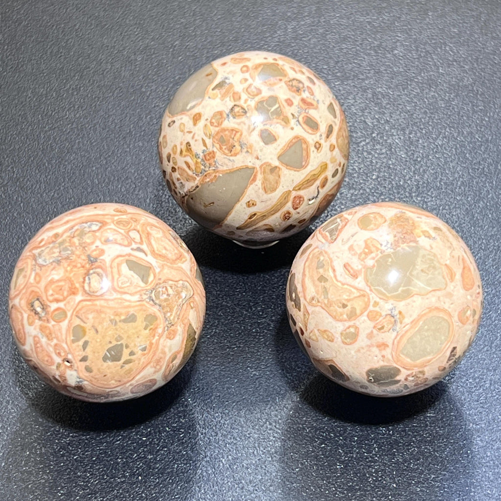 Three polished leopardite spheres showing varied cream, tan, peach, and gray patterns on a dark surface.