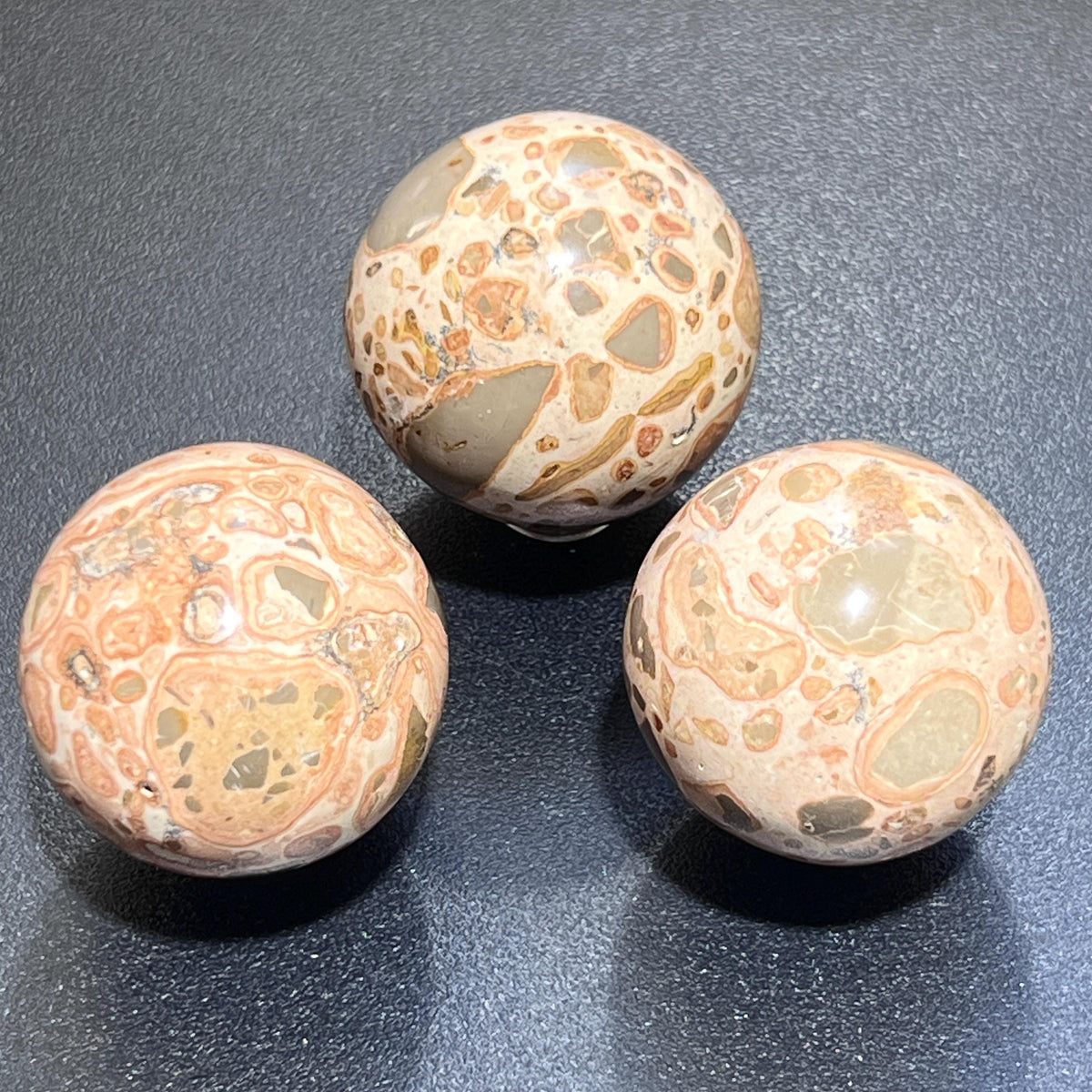 Three polished leopardite spheres showing varied cream, tan, peach, and gray patterns on a dark surface.