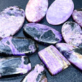 Close-up of polished purple charoite cabochons with lavender swirls and dark markings in assorted oval and rectangular shapes.