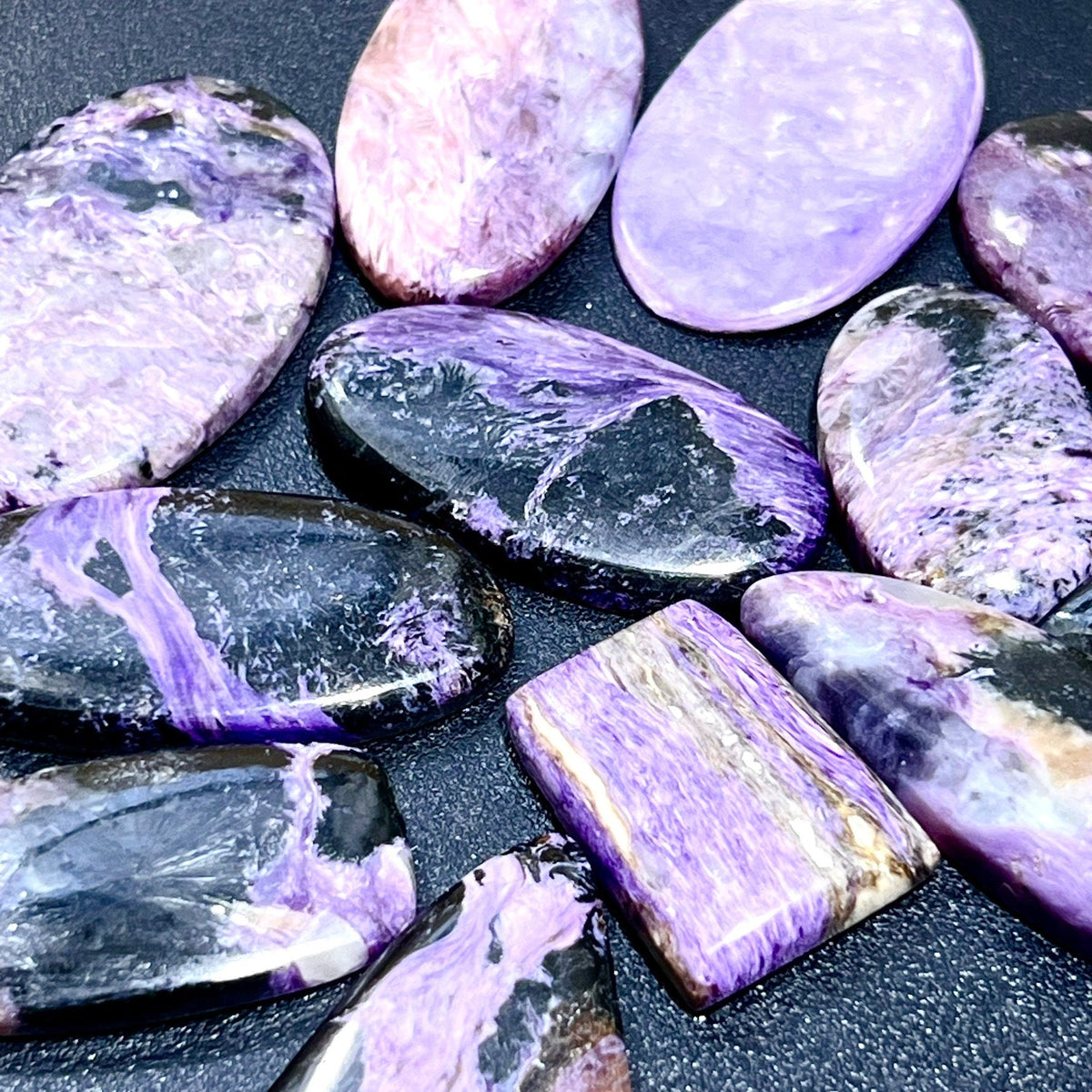 Close-up of polished purple charoite cabochons with lavender swirls and dark markings in assorted oval and rectangular shapes.