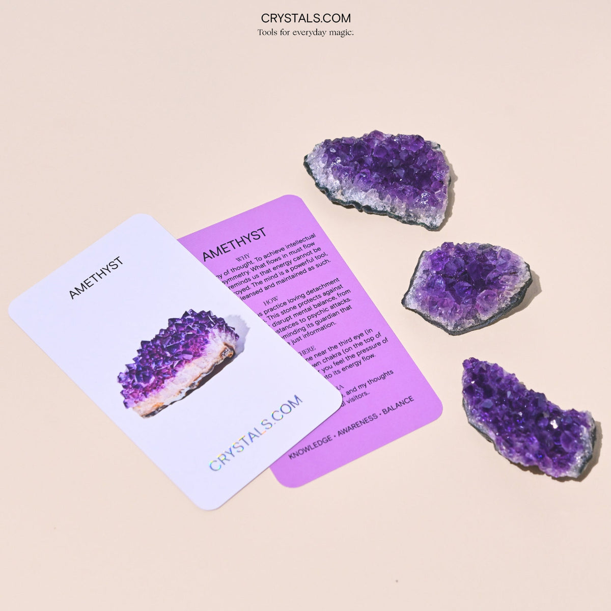 Three purple amethyst clusters beside two Amethyst collector cards on a pale surface.
