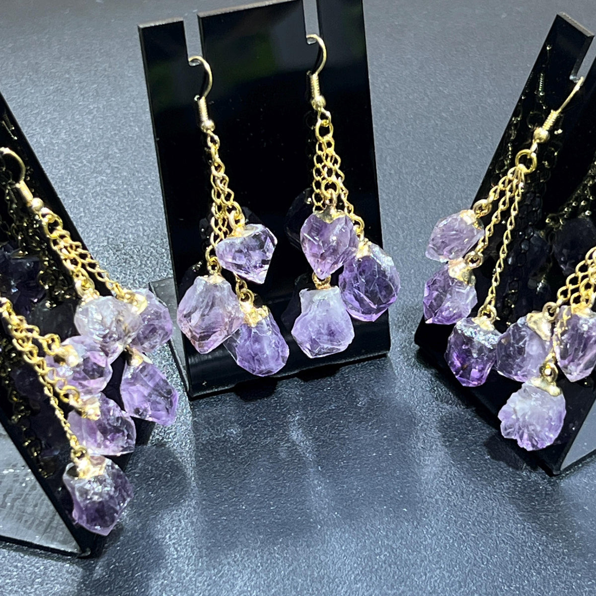 Close-up of purple amethyst crystal drops suspended from staggered gold-tone chains on black earring stands.