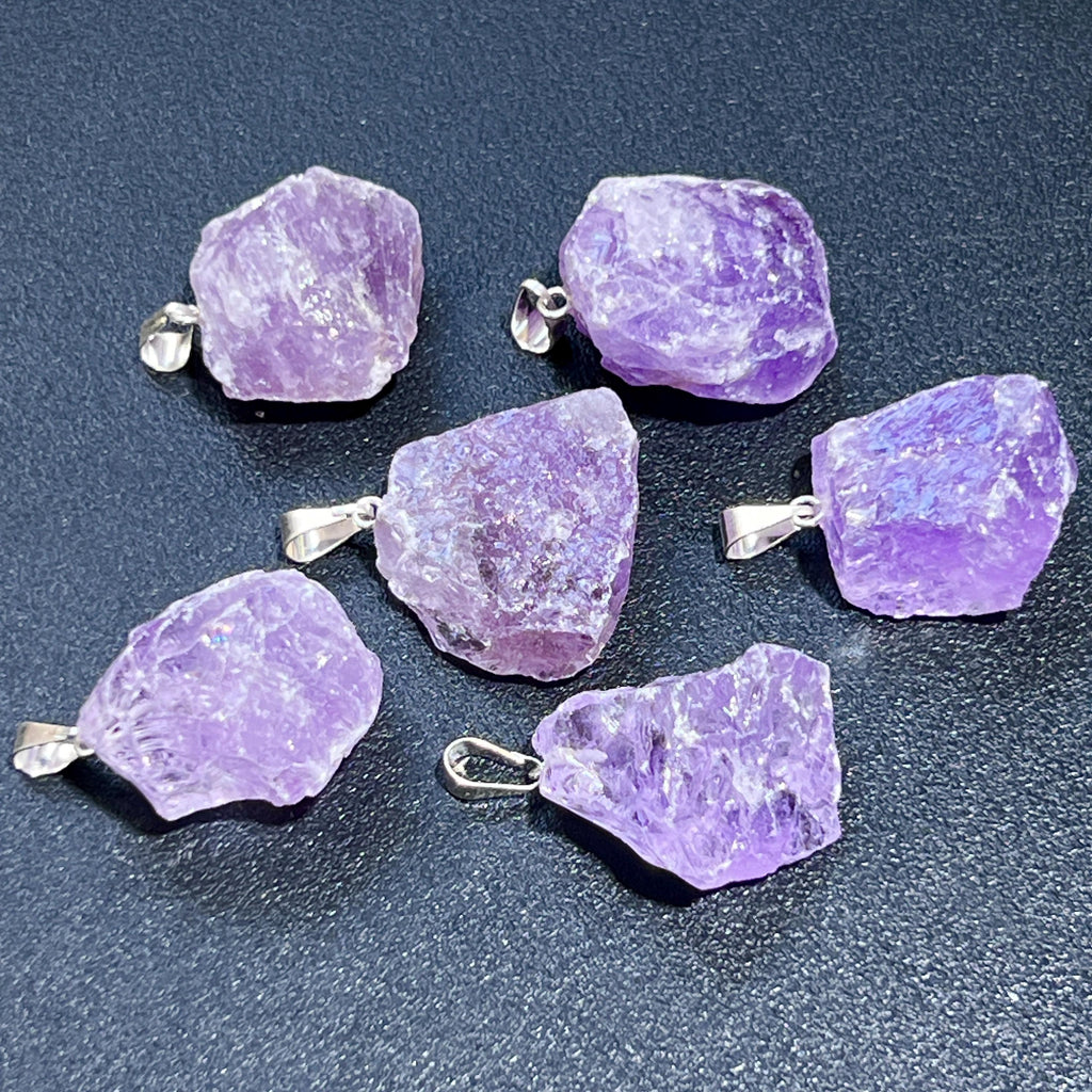 Six rough purple amethyst pendants with silver-tone bails arranged on a dark textured surface.
