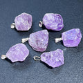 Six rough purple amethyst pendants with silver-tone bails arranged on a dark textured surface.
