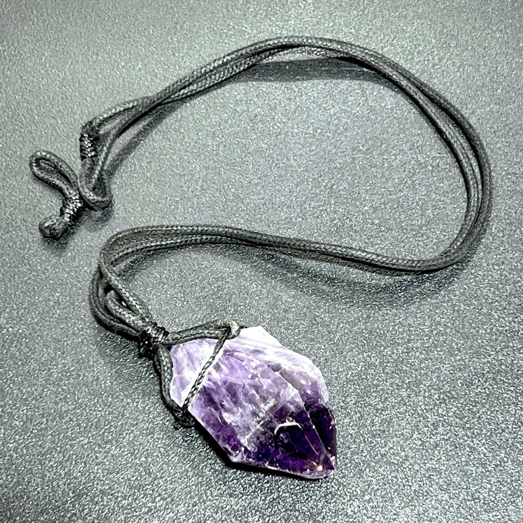 Single raw purple amethyst point pendant with its black cord laid in a loop on a dark surface.