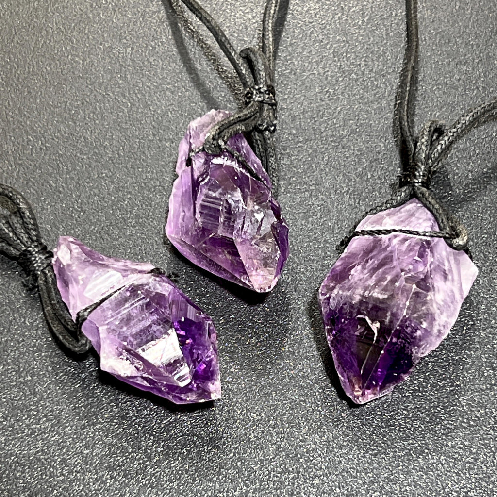Three raw purple amethyst point pendants tied with black cords on a dark textured surface.