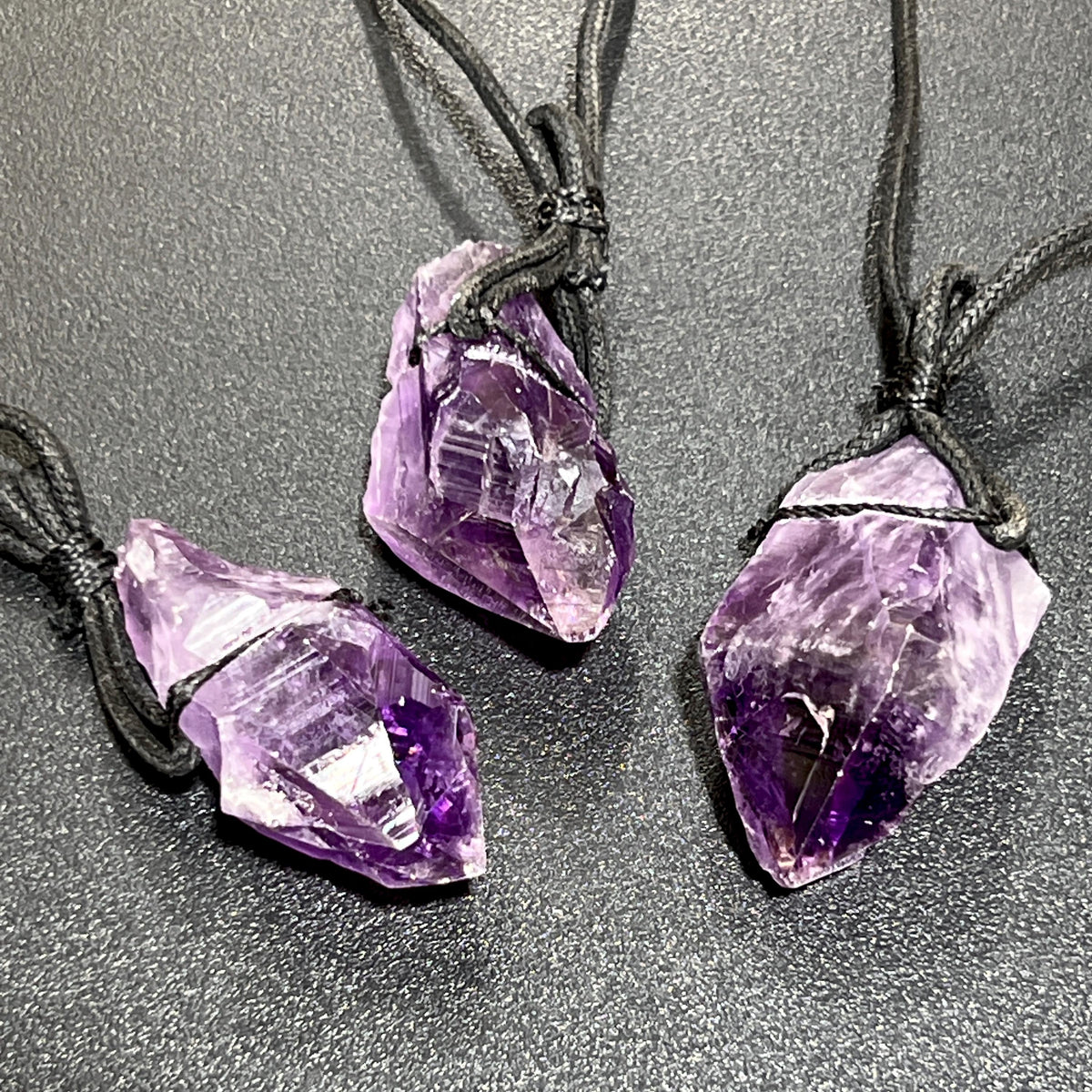Three raw purple amethyst point pendants tied with black cords on a dark textured surface.