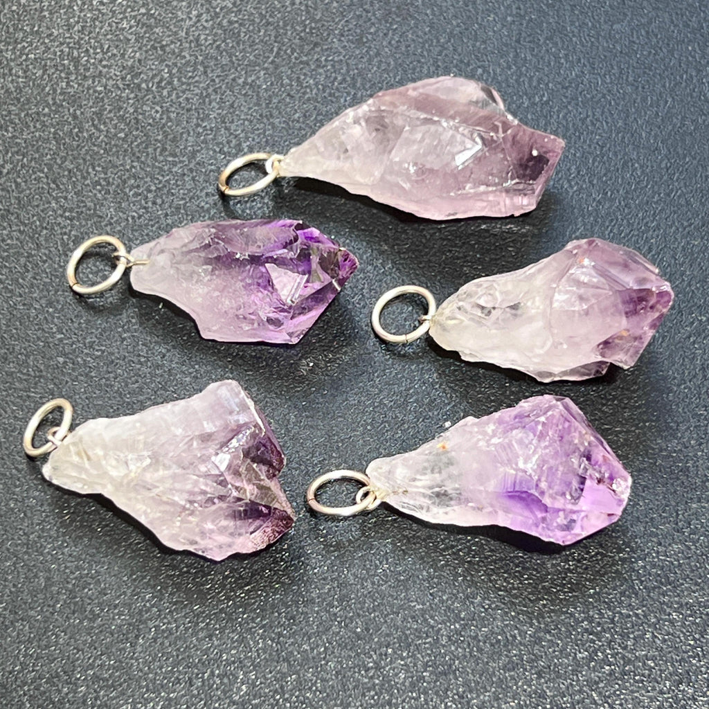 Five raw lavender and purple amethyst point pendants with silver-tone jump rings on a dark textured surface.
