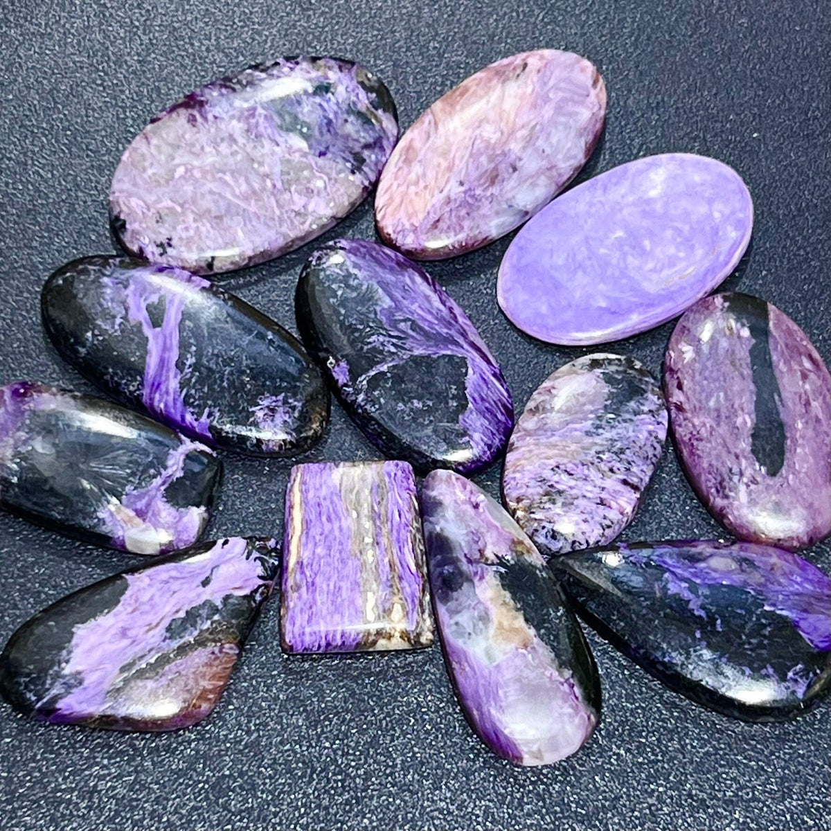 Assorted purple charoite cabochons with pale and dark contrasting patterns arranged on a dark textured surface.