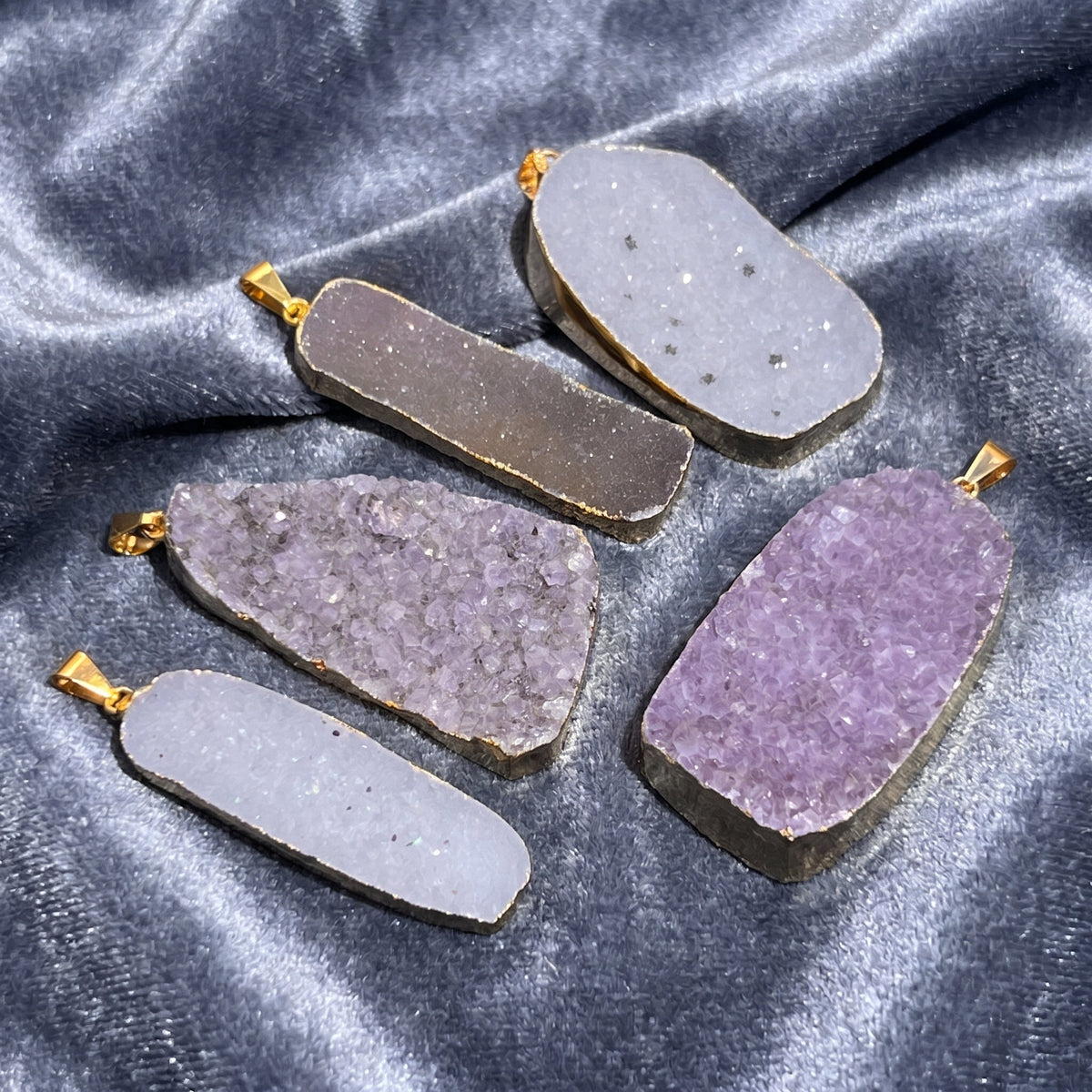 Five assorted pale and purple druzy pendants with gold-plated bails arranged on silver-gray velvet