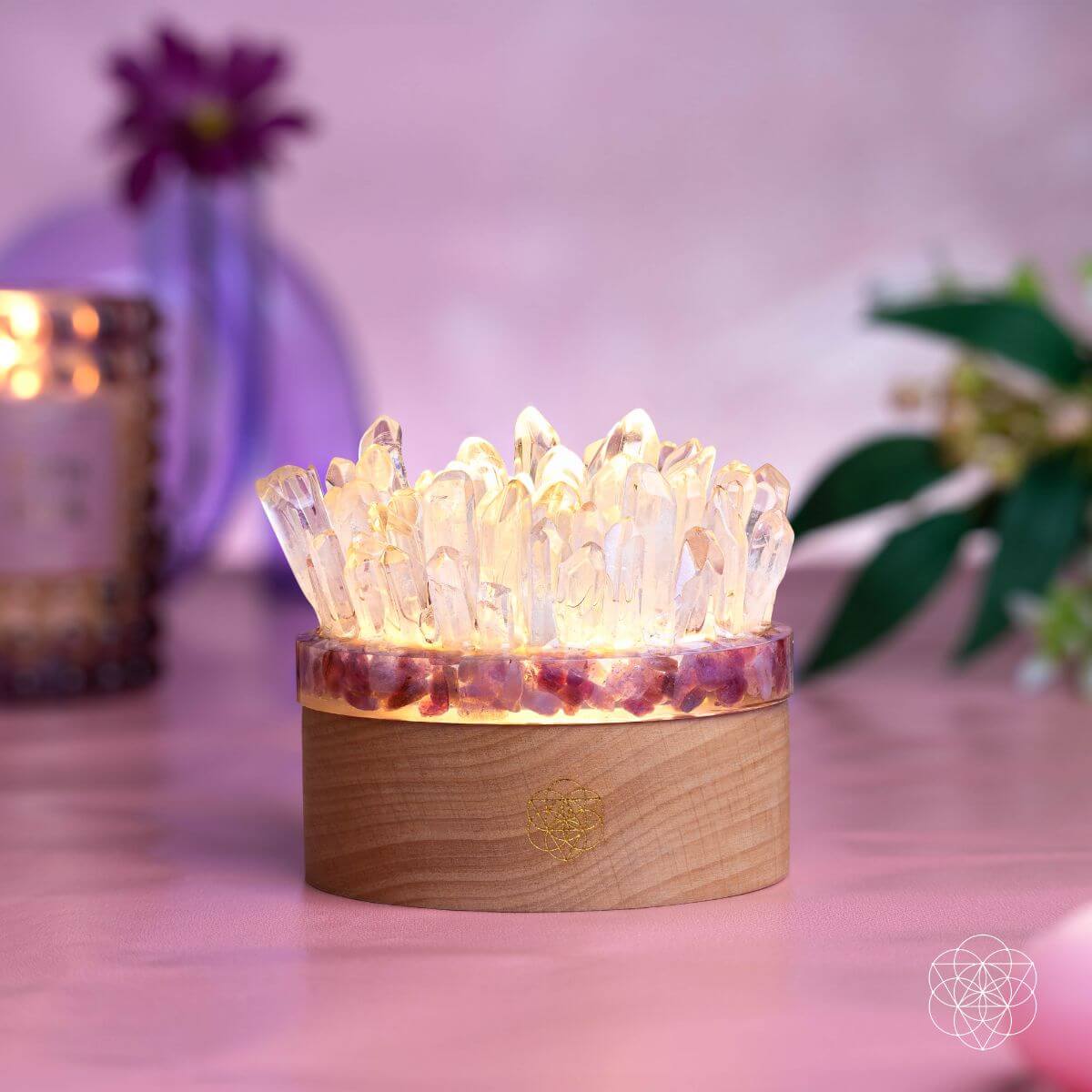 Illuminated quartz and pink tourmaline crystal lamp on a pink tabletop with flowers and a candle behind it.
