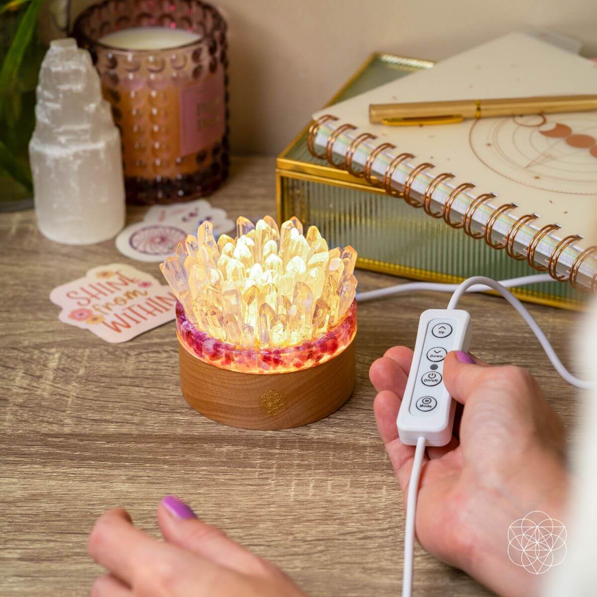 Hand operating the wired inline controls of a glowing quartz and tourmaline lamp beside a notebook.
