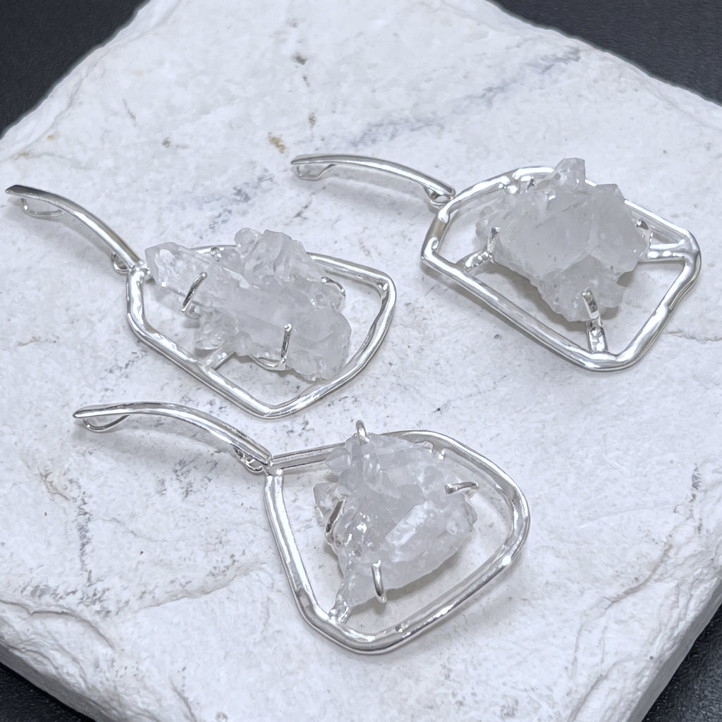 Three quartz cluster statement pendants in silver-plated frames arranged on a white veined stone slab.
