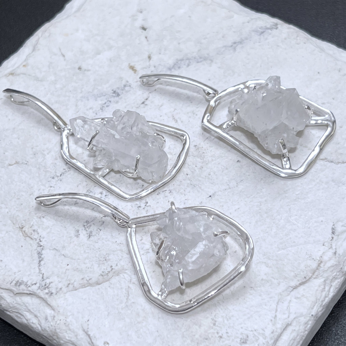 Three quartz cluster statement pendants in silver-plated frames arranged on a white veined stone slab.
