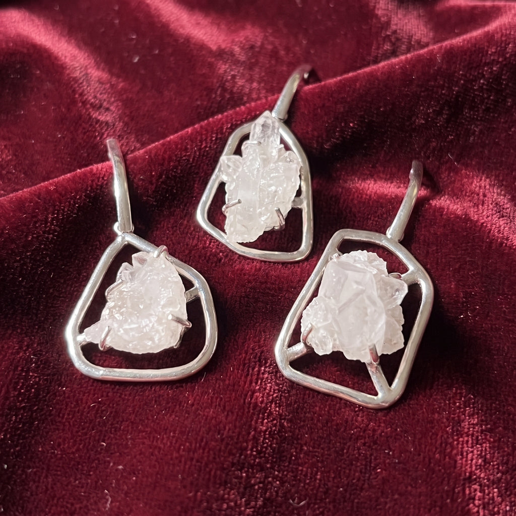 Three quartz crystal cluster pendants in silver-plated geometric frames displayed on burgundy velvet.
