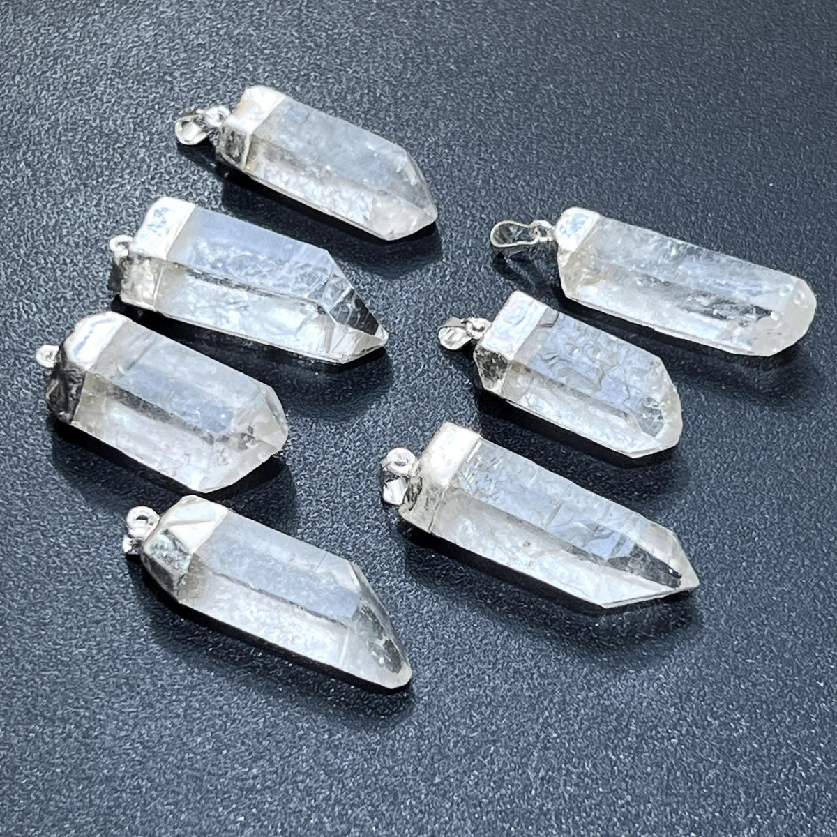 Seven clear quartz crystal point pendants with silver-plated caps and hanging loops on a dark textured surface.
