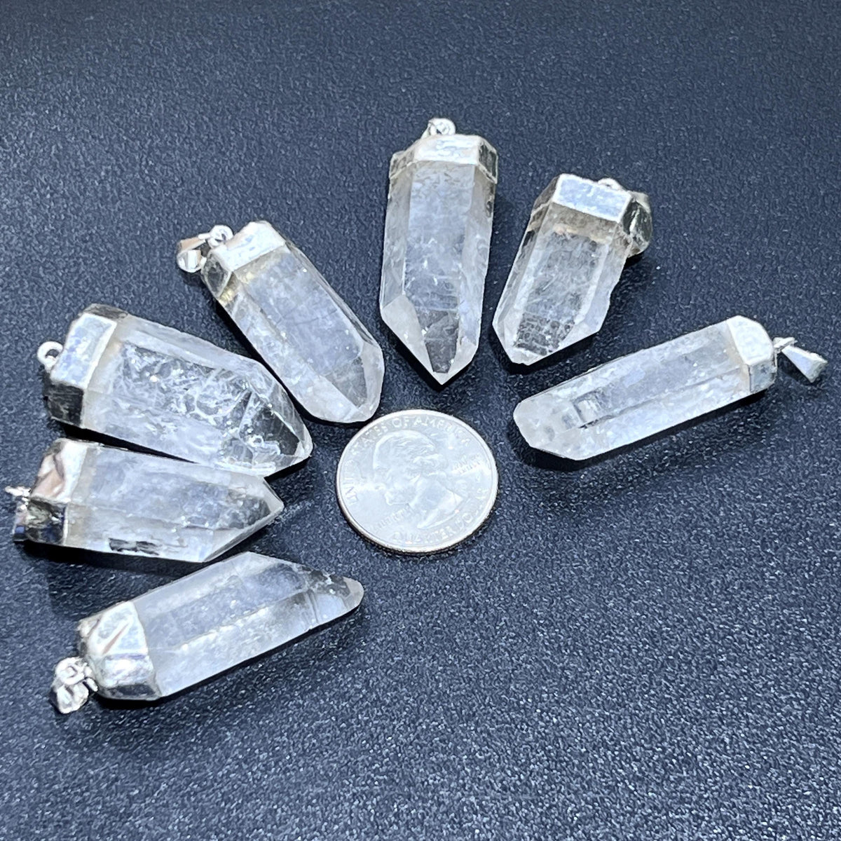 Seven clear to cloudy quartz point pendants with silver-plated caps arranged around a quarter for size comparison.
