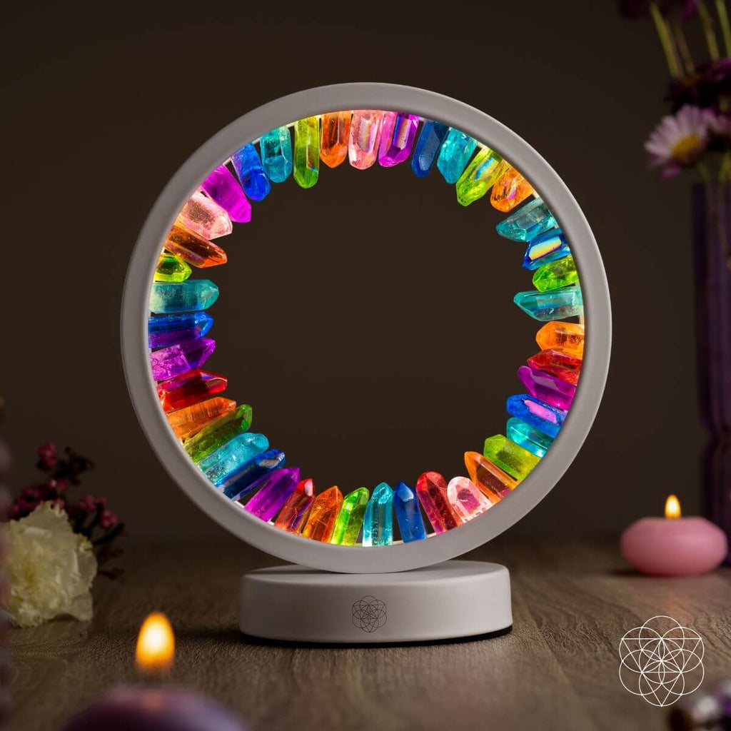 Illuminated rainbow aura quartz halo lamp with a white frame on a wooden table beside candles and flowers.
